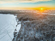 Snow-covered forest on lake shore with ice at sunset and the city on horizon, auerial view