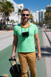 A traveler with camera and luggage on streets of Miami. A man traveler enjoys his summer vacation in a tropical city. A traveler tourist with suitcase on summer city. Man traveler walk on city.