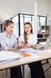 © SOMKID - Two businesswomen discussing work using digital tablet and laptop in modern office, Business team analyzing financial data on a digital tablet and paper report.