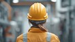 © 69 - Female worker in a yellow hard hat and jacket observed from behind in an industrial setting with machinery and equipment, symbolizing safety and professionalism