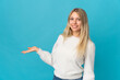 © luismolinero - Young blonde woman isolated on blue background extending hands to the side for inviting to come
