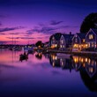© Hutan - Picturesque harbor town at twilight.  Houses with dark gray siding reflect in still water, lit by warm interior lights.  Purple-pink sunset sky.  Small boats on the water