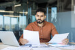 © Liubomir - Young man analyzing financial reports and paperwork, using a laptop, and working diligently in a modern office environment, focusing on professional tasks