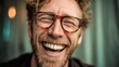 © mila103 - A cheerful man with curly hair and a beard is laughing with a big smile. He wears stylish glasses and is sitting in a bright cozy indoor setting radiating positivity.