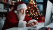 © Alexander - Santa Claus in red costume working at office desk with modern computer, amazed by digital letters and wish lists, Christmas tree with lights in background. santa claus office, red
