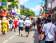 © ozguryaman - Blurred, defocused crowd of unrecognizable people walking down a busy street lined with shops and tropical greenery under a sunny blue sky. Captures the vibrant atmosphere of a summer street festival