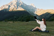 © SHOTPRIME STUDIO - A woman sits on a small bag in a grassy field, using a laptop with a mountain backdrop. She appears focused and creative, conveying a productive outdoor work scene.