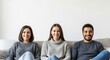© Николай Срибяник - Three diverse friends (two women and one man, mixed ethnicity) in casual sweaters and jeans sit smiling happily side-by-side on a light sofa against a white background.