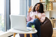 © Sofiia Shunkina - Young adult woman sitting on beanbag holding smartphone gesturing OK sign. Cozy home office environment with laptop, bright window, bookshelf. Concept of confidence work-life balance positivity