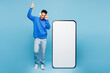 © ViDi Studio - Full body young man of African American ethnicity wear sweatshirt casual clothes headphones big huge blank screen mobile cell phone smartphone with area listen music isolated on plain blue background