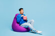 © ViDi Studio - Full body young man of African American ethnicity wear sweatshirt casual clothes sit in bag chair hold takeaway bucket eat popcorn watch movie film isolated on plain pastel light blue cyan background.