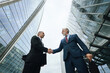 © DragonImages - Upward view of two middle aged Caucasian men shaking hands outdoors between modern glass skyscrapers, both wearing business attire, engaging in professional agreement or partnership
