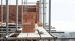 © Ulum - Close-up of a brick wall under construction with scaffolding, showing bricks, mortar, and a trowel on a wooden platform.
