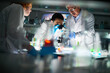 © pressmaster - Multi-ethnic group of chemists doing research in modern laboratory. Pretty young woman in protective mask looking through microscope while her coworkers standing nearby