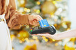 © Alliance - A close-up shows womans hand making contactless credit card payment at point-of-sale terminal. Blurred festive lights in background suggest modern holiday shopping convenience and quick transaction