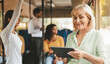 © Prostock-studio - A woman smiles while using a tablet on a bus. She is surrounded by other passengers, including a person standing and others sitting.