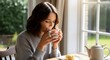 © Fdl Studio99 - A serene woman sips a warm drink from a mug while sitting at a sunlit table indoors with a teapot and pastry.