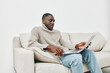 © SHOTPRIME STUDIO - Young African man in cozy sweater working from home on laptop, showcasing productivity and relaxation in a modern living room