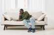 © SHOTPRIME STUDIO - Young Black man sitting on a cozy sofa, working on a laptop with a focused expression, wearing a green sweater and casual jeans in a bright, modern living room
