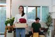 © itchaznong - Moving day. Young woman carrying a box of clothes with a smile in a bright living room.