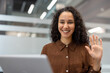 © Liubomir - Young mixed-race woman smiling and waving at camera during an online video call on her laptop from a modern office, friendly and professional remote work connection
