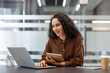 © Liubomir - Professional young woman with curly hair working on a laptop at a modern office desk, holding a notebook and pen while smiling, expressing engagement and productivity