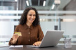 © Liubomir - Smiling woman holding a credit card while typing on a laptop, making an online purchase or payment in a modern office setup, concentrating on the transaction