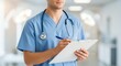 © Alice O - Medical professional in blue scrubs holds clipboard and pen while wearing stethoscope in modern hospital. Clean, bright healthcare environment with blurred background creates atmosphere.