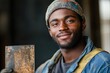 © DesignMaster360 - A young, smiling Black man in a beanie and work clothes holds a rusty metal plate, suggesting craftsmanship or a blue-collar profession.
