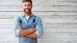 © GrayFrog - Smiling handyman with a beard, denim shirt and overalls, standing with crossed arms in front of a wooden wall.