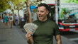 © Krakenimages.com - Young man holding us dollars on an urban street with a confident smile amid bustling city life and public transportation in the background