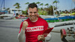 © Krakenimages.com - Man wearing red volunteer shirt standing beachfront holding clipboard showing thumbs up confident expression amid palm trees and boats suggesting outdoor community service involvement.