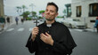 © Krakenimages.com - Priest holding cross while standing on urban street with trees and buildings in the background, man in religious attire looks contemplative outdoors in city environment.