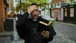© Krakenimages.com - Priest reading book on urban street with shocked expression wearing black robe and cross necklace with blurred cityscape background showing trees and buildings