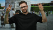 Young man with beard making peace signs in front of pisa tower in italy, wearing glasses outdoors with crowd in background, showcasing travel and tourism vibe