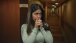 © Krakenimages.com - Young hispanic woman clasping hands near mouth with wide eyes in hotel building; anxiety tension worry fear.