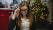© Krakenimages.com - Young woman uses hand to adjust glasses on street outside building decorated with lights and garlands; confidence.