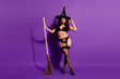 © deagreez - Glamorous witch in black lingerie with broom stands against purple backdrop portraying Halloween charm and elegance