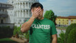 Asian man wearing green volunteer shirt standing in front of pisa tower in italy, expressing a thoughtful emotion in the outdoor setting, with iconic landmark in background.