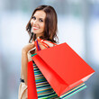© vgstudio - Happy smile young cute woman in dress carrying colorful shopping packages bags, stand against blurred mall background. Brunette girl in black Friday sales discounts offers ad concept. Square image