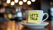 © Design Stock - A white coffee cup with a bright yellow sticky note that says lets talk attached to it set on a saucer with a blurred background of warm lights