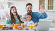 © InsideCreativeHouse - Family with little son sharing morning meal around the kitchen table enjoying tasty food drinking orange juice have video call on laptop waving hands , eating breakfast together in modern apartment.