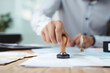 © Starmarpro - Close up of a hand using a stamp on documents at a desk, representing approval, authentication, or processing paperwork.