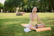 © m-art - A young woman sits on a yoga mat in a park, wearing a yellow outfit. She is smiling while talking on her phone, surrounded by green grass and trees.
