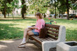 © m-art - A young woman sits on a bench in a green park, absorbed in a book. Her pink jacket brightens her casual outfit as the afternoon sun shines down. A scooter rests nearby.