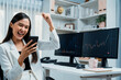© InfiniteFlow - Successful young Asian businesswoman earning high profit raising fist up, looking smartphone for exchange stock market's financial technology with dynamic business graph at modern office. Stratagem.
