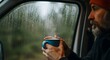 © Rumah Kantor Apart - Cozy Escape in the Rain: A bearded man in a red beanie holds a steaming mug of coffee inside a vehicle, looking out at the rain-streaked window as he finds comfort and solitude amidst a blurred.