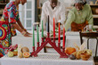 © AnnaStills - Three Black adults celebrating Kwanzaa, arranging festive table with kinara holding seven lit candles, assorted fruits, vegetables, and traditional decorations in foreground