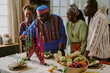 © AnnaStills - Group of Black adults and young adult woman celebrating Kwanzaa, lighting kinara candles together at table with traditional fruits and decorations, smiling and interacting