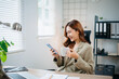 © laddawan - Asian businesswoman holding coffee cup and smartphone in bright office. Perfect for business lifestyle, communication, productivity, and modern workplace themes.